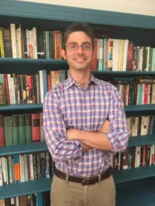 Jeffrey Gonzalez stands in front of a bookshelf with his arms crossed. He is wearing glasses and a blue and white plaid dress shirt. 