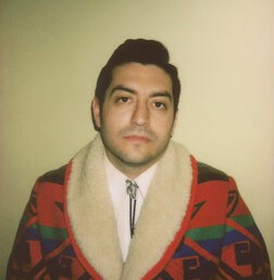 A man stands solemnly in front of a plain tan wall wearing a brightly colored coat with a southwestern pattern over a white collared shirt and a bolo tie